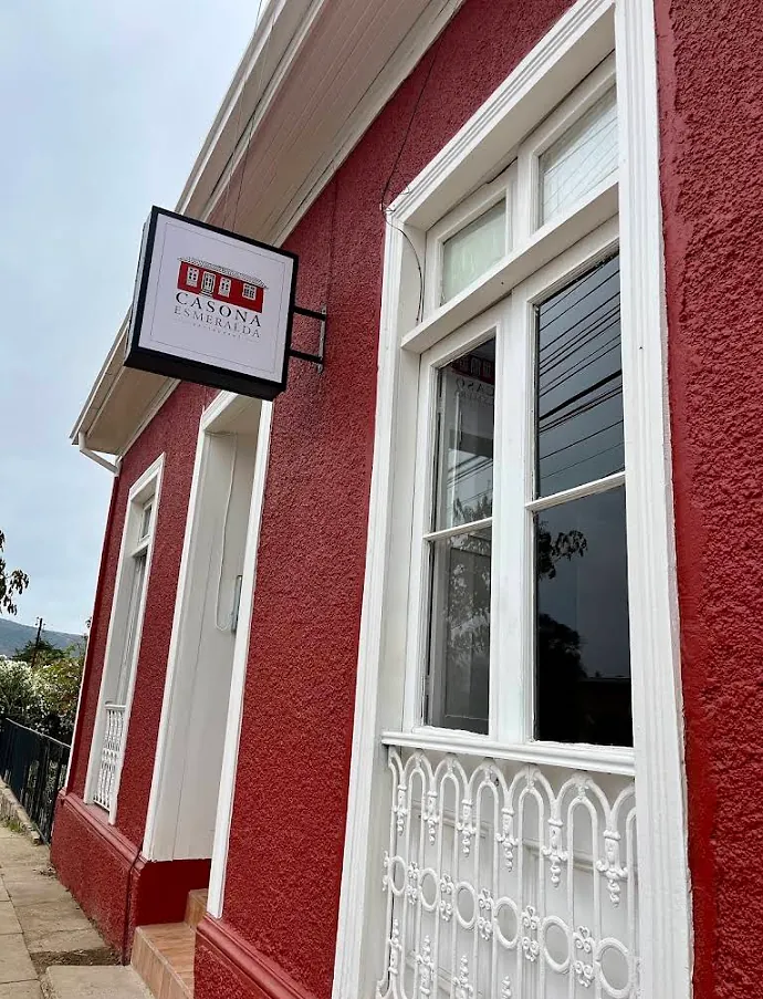 Fachada de Casona Esmeralda a la luz del día, con muros rojos, marcos blancos, reja forjada y el letrero con el logo