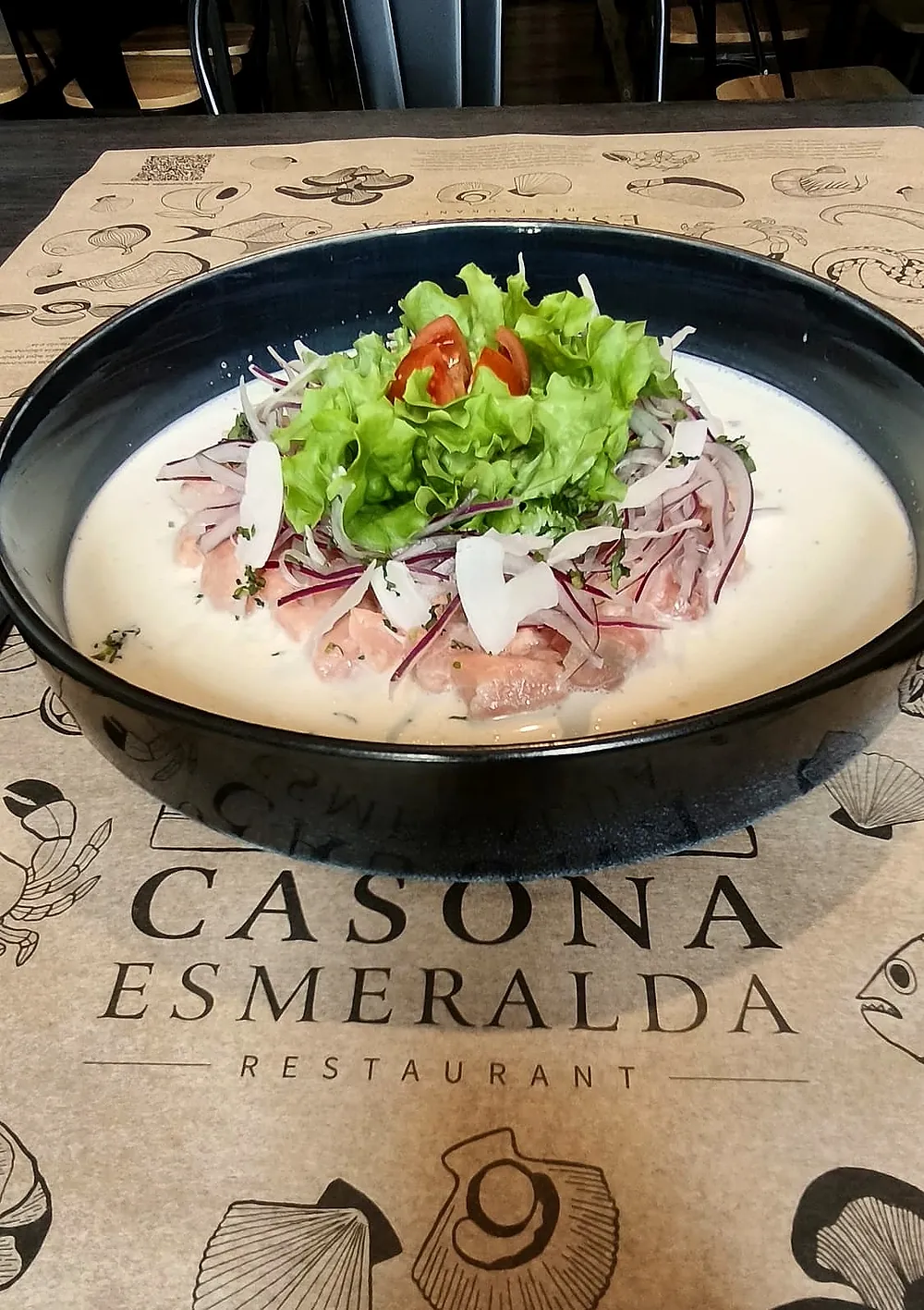 Ceviche en un plato hondo negro sobre un individual de papel con el logo del restaurante