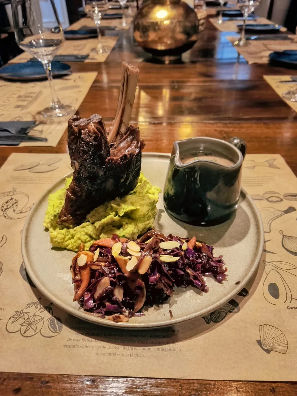 Plato de carne con hueso sobre puré verde y repollo morado, junto a una jarra de salsa